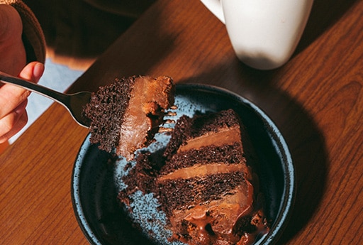 Someone sitting at a table with a cup of coffee, scooping a chocolate cake from a small plate.